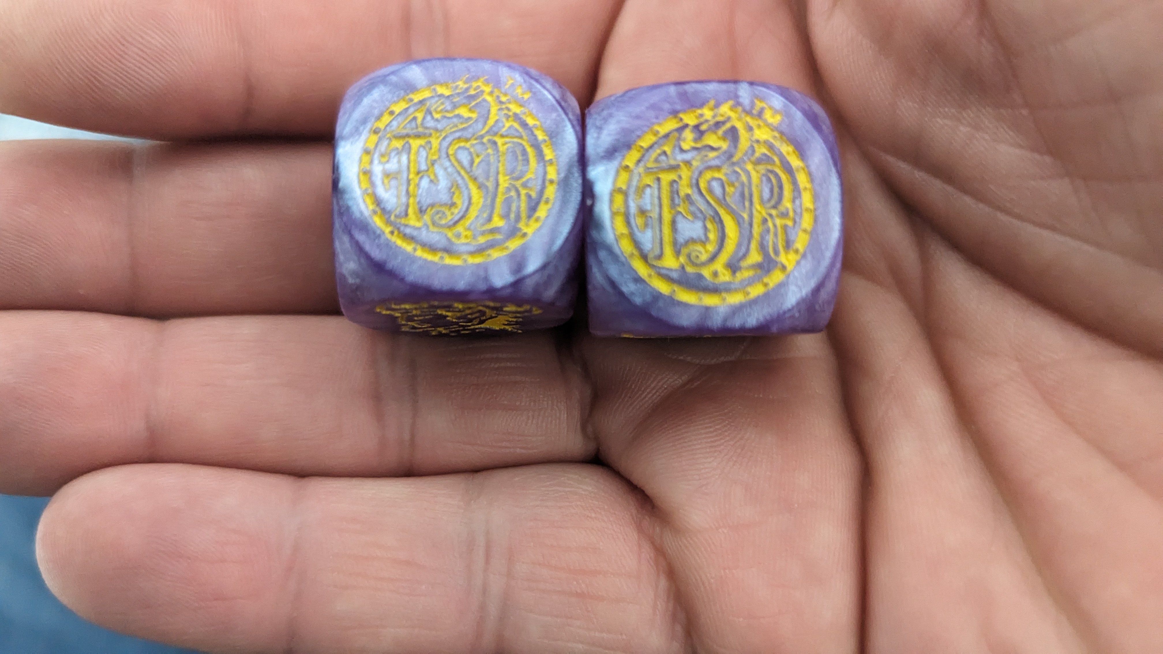 Two purple cubic dice with the TSR dragon logo displayed on a white man’s hand.